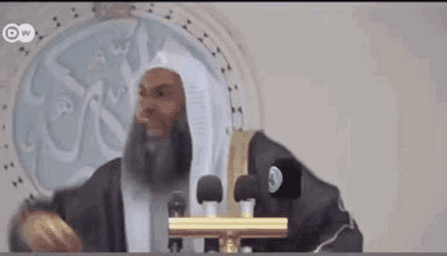 a man with a beard is giving a speech in front of two microphones in a mosque .