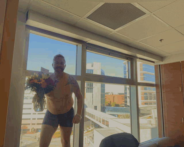 a shirtless man holding a vase of flowers stands in front of a window