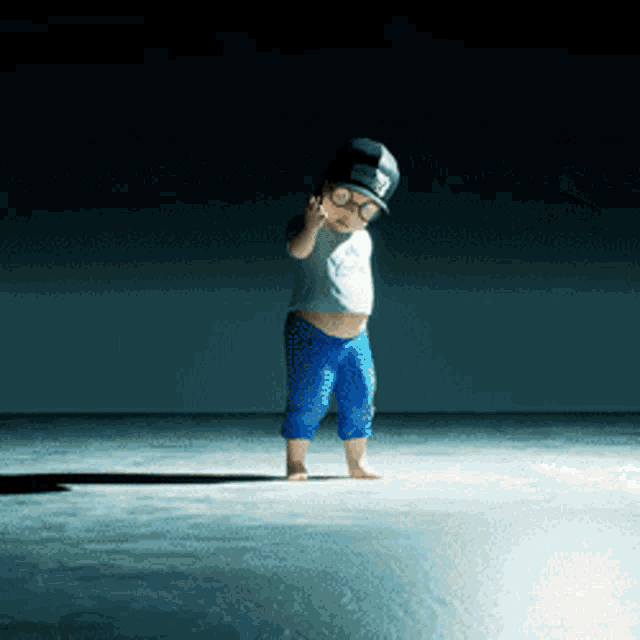 a little boy wearing a hat and glasses stands on a concrete floor