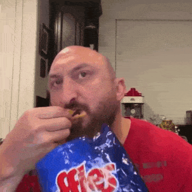 a bald man with a beard is eating a bag of fritos