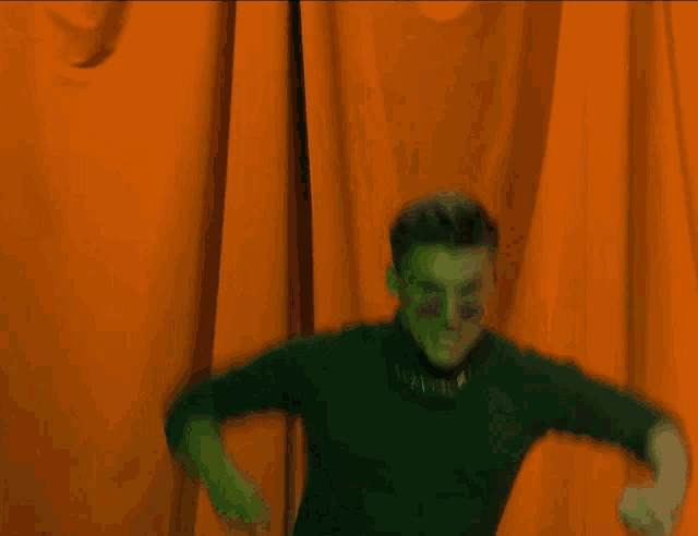 a man wearing sunglasses and a green turtleneck stands in front of a orange curtain