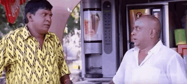 two men are standing next to each other in front of a vending machine and talking to each other .