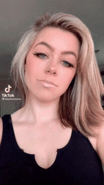 a woman with blonde hair is wearing a black tank top and has a tiktok sticker on her face