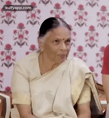 an elderly woman in a white saree is sitting in front of a wall with a pattern .
