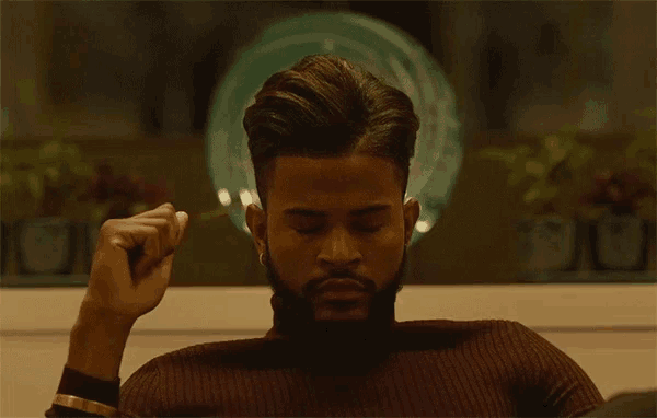 a man with a beard wearing a brown turtleneck sweater holds his fist up