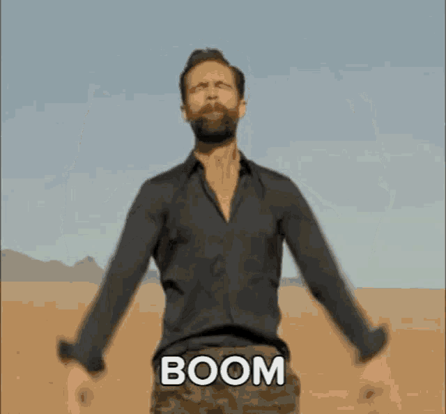 a man with a beard is standing in the desert with his arms outstretched and the word boom written on his pants .
