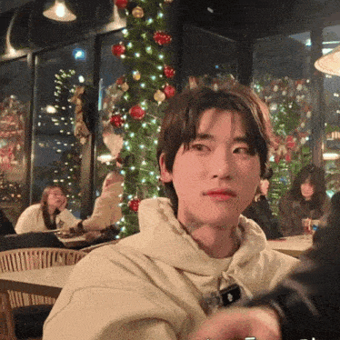 a young man with a tattoo on his arm looks at the camera in front of a christmas tree