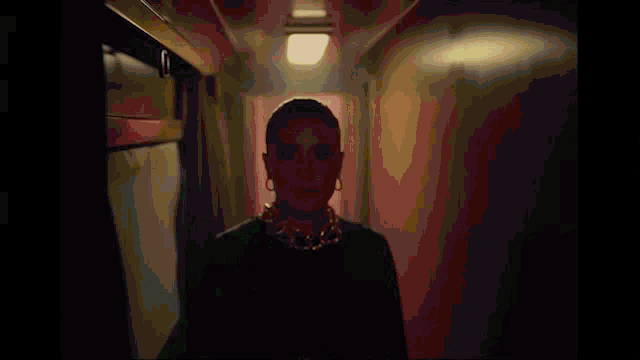 a woman wearing a black dress and a gold chain around her neck is standing in a hallway .
