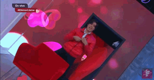 a man in a red jacket is standing in front of a pink heart on a television screen