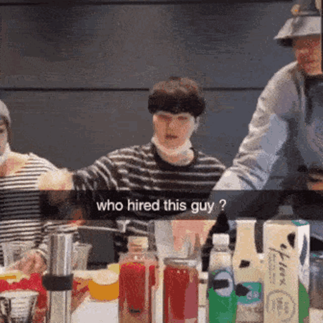 a group of people are sitting around a table with bottles of drinks and a man is asking who hired this guy .