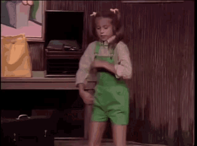a young girl in green overalls is dancing in front of a record player .