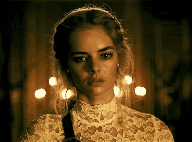 a woman wearing a white lace top has a tear running down her face