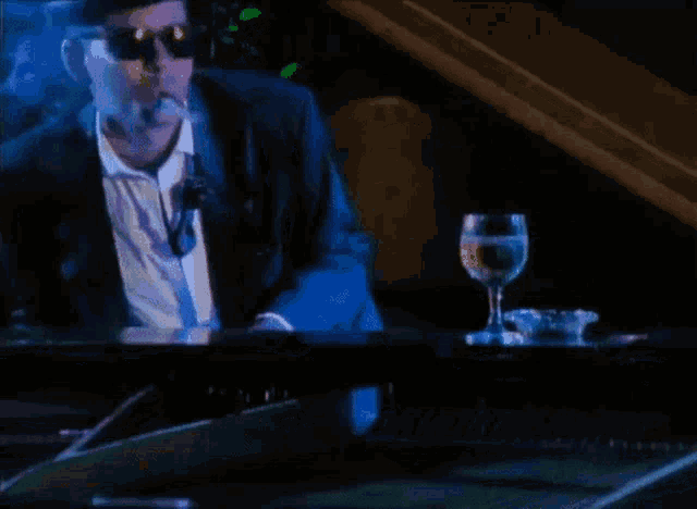 a man in a suit is smoking a cigarette while playing a piano next to a glass of wine .