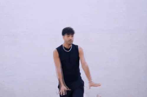 a man wearing a black tank top and black pants is dancing in front of a white wall