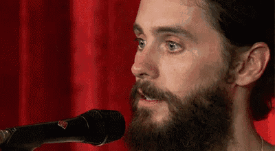 a man with a beard is singing into a microphone with a red curtain behind him .