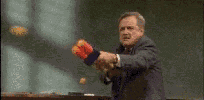a man in a suit is holding a nerf gun in front of a blackboard in a classroom .