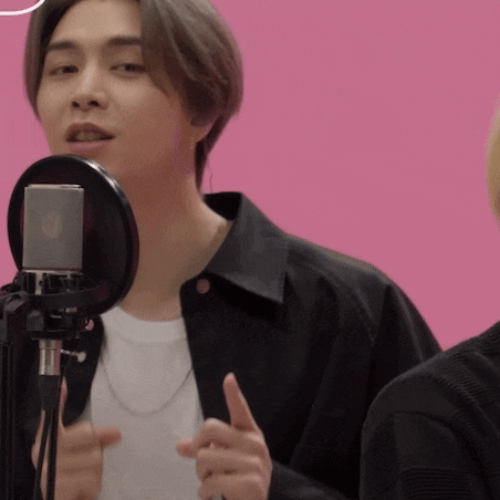 a man in a black jacket is singing into a microphone on a pink background