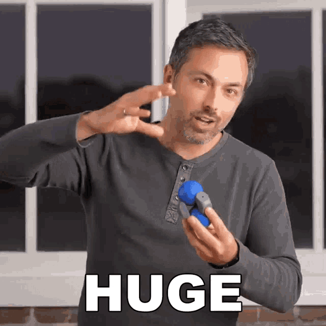 a man in a gray shirt holds a blue object and says " huge " in white letters