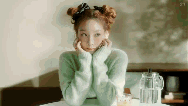 a woman in a green sweater is sitting at a table with her hands on her face and a pitcher of water .