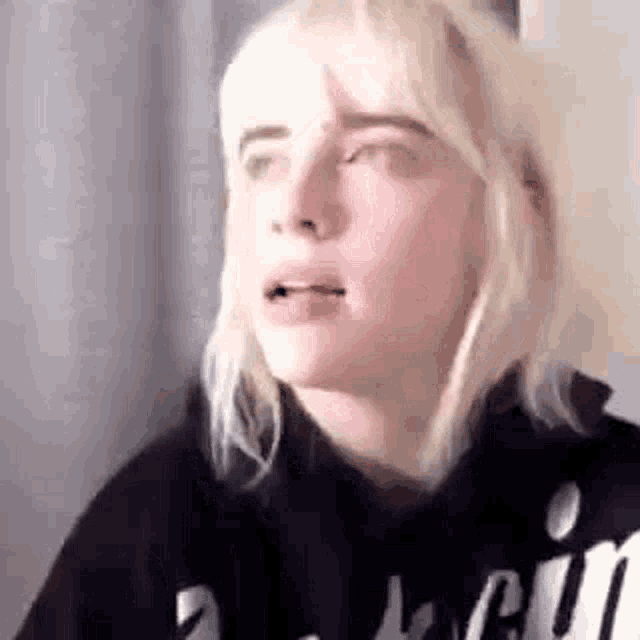 billie eilish is wearing a black hoodie and making a face .