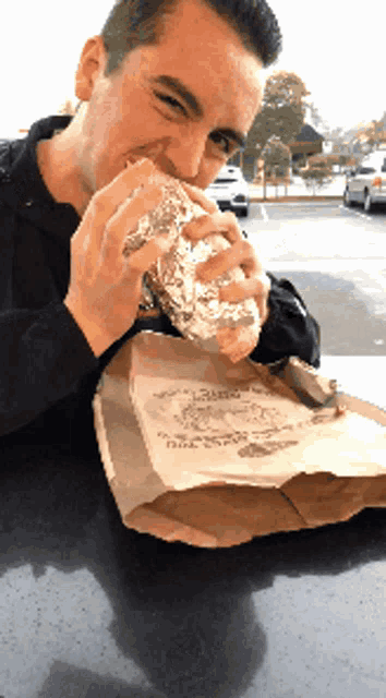a man sitting at a table eating a sandwich from a bag that says no toppings