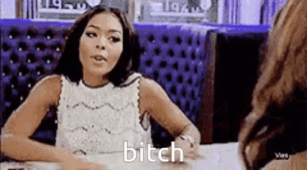 a woman in a white dress is sitting at a table in a diner talking to another woman and says bitch .