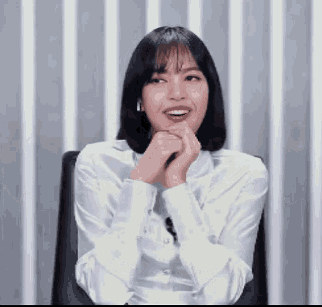 a woman in a white shirt is sitting in a chair with her hands on her chin and smiling .
