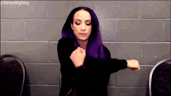 a woman with purple hair is sitting in a chair with her arms outstretched in front of a brick wall .