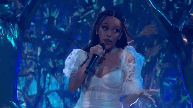 a woman in a white dress is singing into a microphone while standing in front of a blue background .