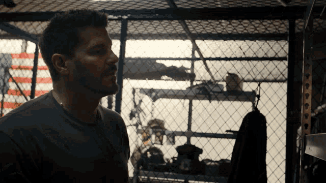 a man with a beard stands behind a chain link fence in a dark room