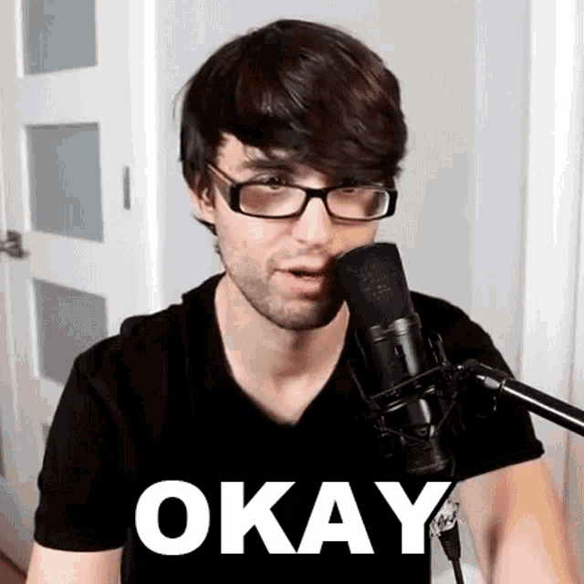 a man wearing glasses is talking into a microphone and the word okay is on the bottom of his shirt .