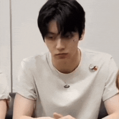 a young man wearing a white t-shirt is sitting at a table with his hands folded .