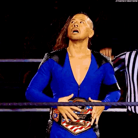 a wrestler in a blue suit is holding an american flag belt while standing in a ring .