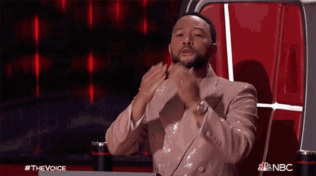 a man in a suit is sitting in a chair with his hands in his pockets and a nbc logo in the background .