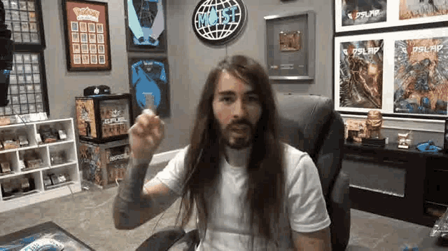 a man with long hair and a beard is sitting in a chair in a room with a lot of posters on the wall .