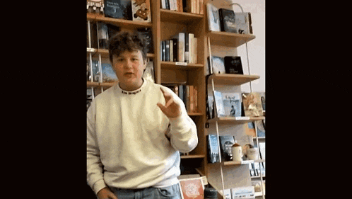 a man in a white sweater is standing in front of a bookshelf in a bookstore .