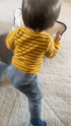 a baby in a yellow striped shirt and blue pants is standing on a bed .