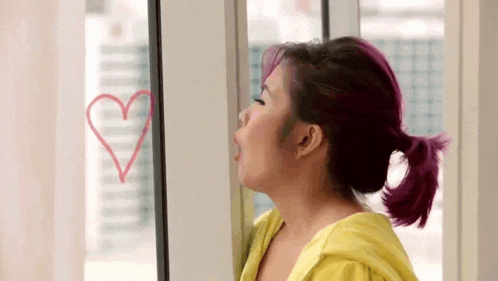 a woman with purple hair is blowing a kiss on a window with a heart drawn on it .