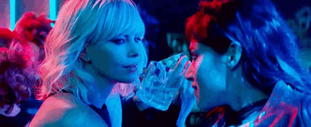 two women are standing next to each other in a dark room . one of the women is drinking from a glass .