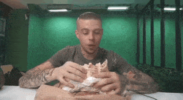 a man with tattoos is sitting at a table eating a hamburger .
