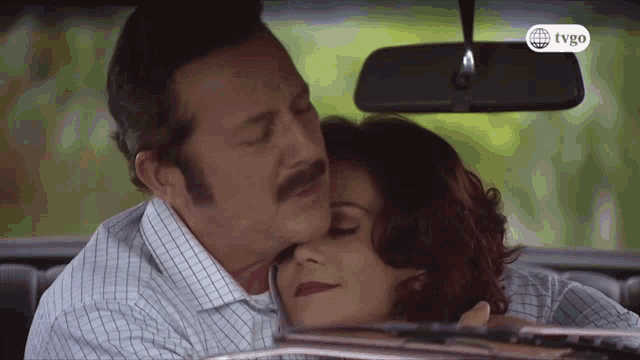a man kisses a woman on the forehead while sitting in a car with a tvgo logo on the rear view mirror
