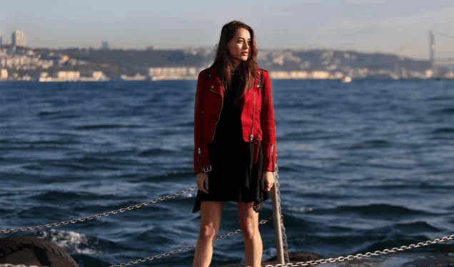 a woman in a red jacket and black dress is standing on a pier overlooking the ocean