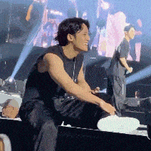 a man in a black tank top is sitting on a stage with a microphone in his hand .