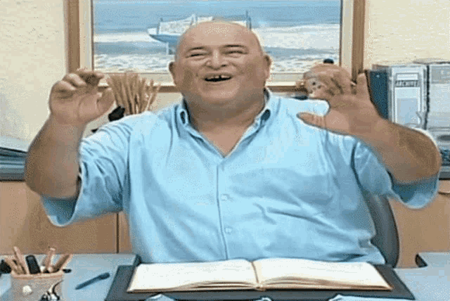 a bald man in a blue shirt is sitting at a desk with an open book on it