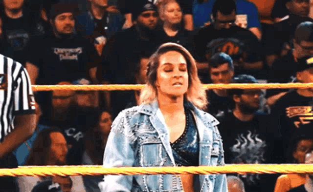 a woman in a denim jacket stands in a wrestling ring with a referee in the background