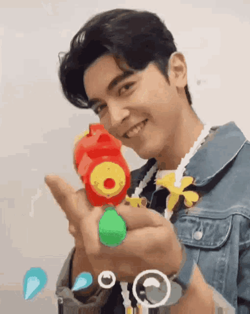a young man in a denim jacket is holding a toy water gun and smiling .