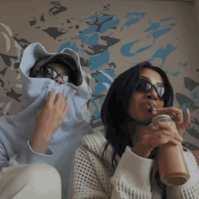 a woman drinking a smoothie with a straw while another woman covers her face with a hoodie