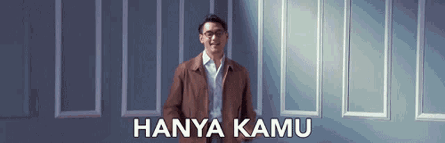 a man in a brown jacket is standing in front of a wall with the words hanya kamu on it