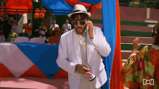 a man in a white suit talking on a cell phone with a woman in a floral dress behind him