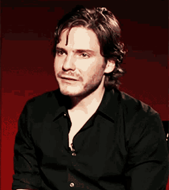 a man wearing a black shirt is sitting in front of a red wall
